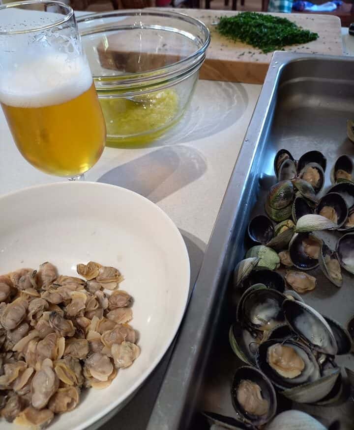Clam meat in a bowl on a bench, an oven tray with steamed clams next to it. A beer glass and a chopping board with chopped herbs in the back ground