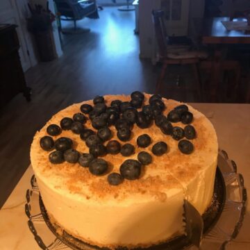 A cheesecake topped with blueberries on a glass platter standing on a white table. A cake serving cutter slicing into the cake