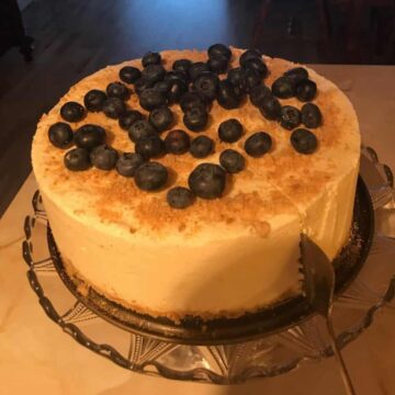 A cheesecake topped with blueberries on a glass platter standing on a white table. A cake serving cutter slicing into the cake