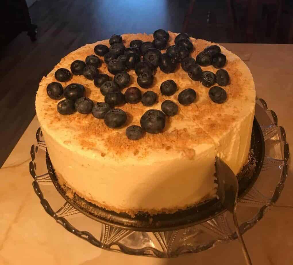 A cheesecake topped with blueberries on a glass platter standing on a white table. A cake serving cutter slicing into the cake