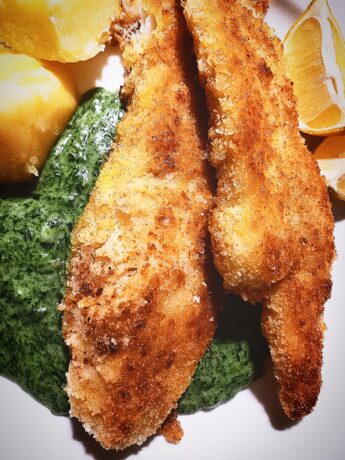 Golden brown crumbed fish fillets served on top of creamed nettles. Boiled potatoes and lemon wedges on the side. All on a white plate