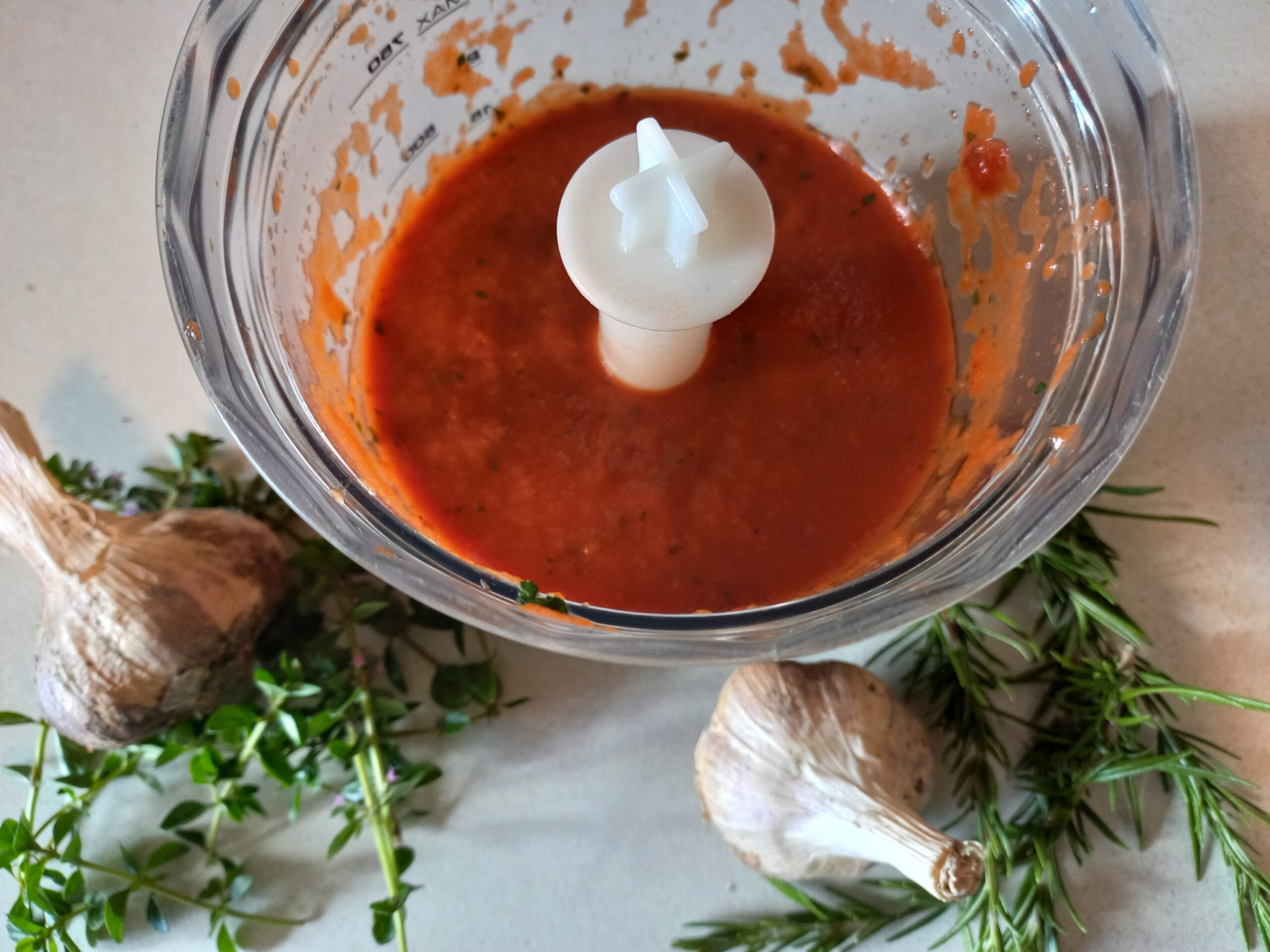Pizza sauce in a food processor bowl. 2 garlic heads, fresh rosemary and thyme on the bench next to it