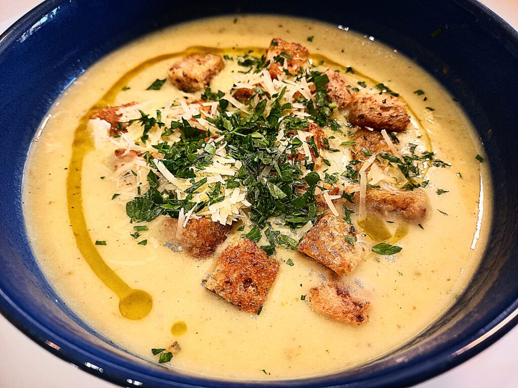 Close up of a blue bowl with roasted garlic and cream soup topped with croutons, grated Parmesan cheese and chopped parsley