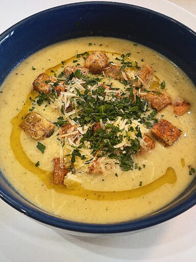 Roasted garlic cream soup topped with croutons, grated Parmesan cheese, chopped parsley and a drizzle of truffle oil. In a blue bowl on a white plate