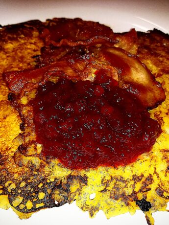 Two Raggmunkar on a white plate topped with crispy bacon and lingon berry jam