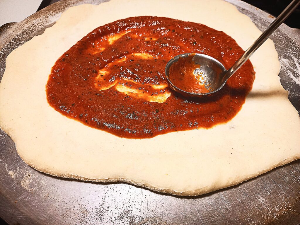 Uncooked pizza base on a pizza tray. A ladle of pizza sauce being spread on top