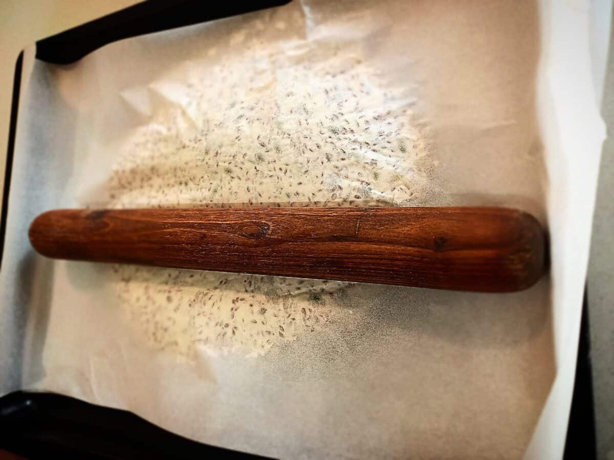 Sunflower seed crisp bread dough on an oven tray topped with parchment paper and a rolling pin on top.