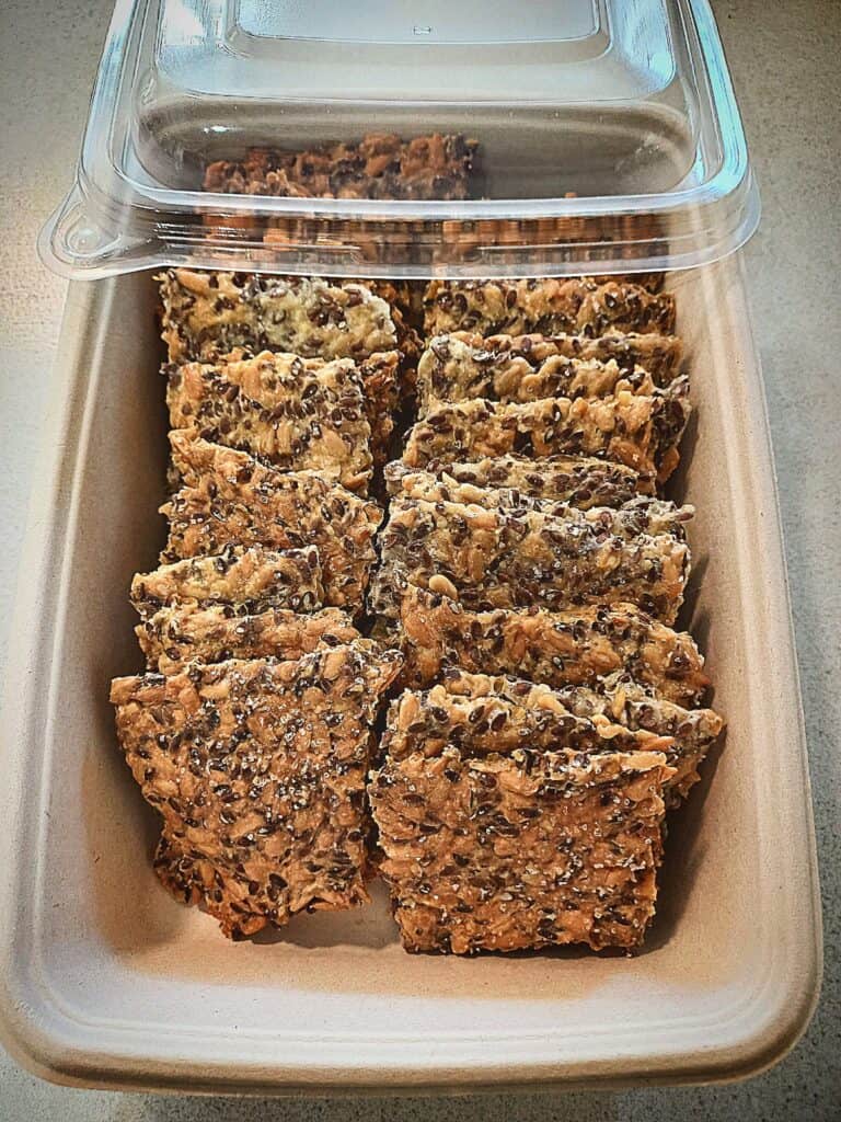 Sunflower seed crisp bread squares in a compostable tray with a plastic lid covering half the product