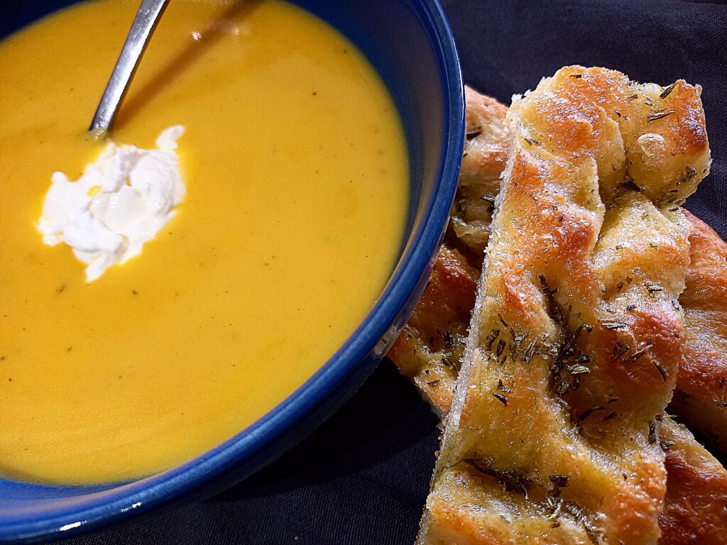 A bowl of pumpkin soup with sour cream in the middle. Sliced focaccia bread on the side