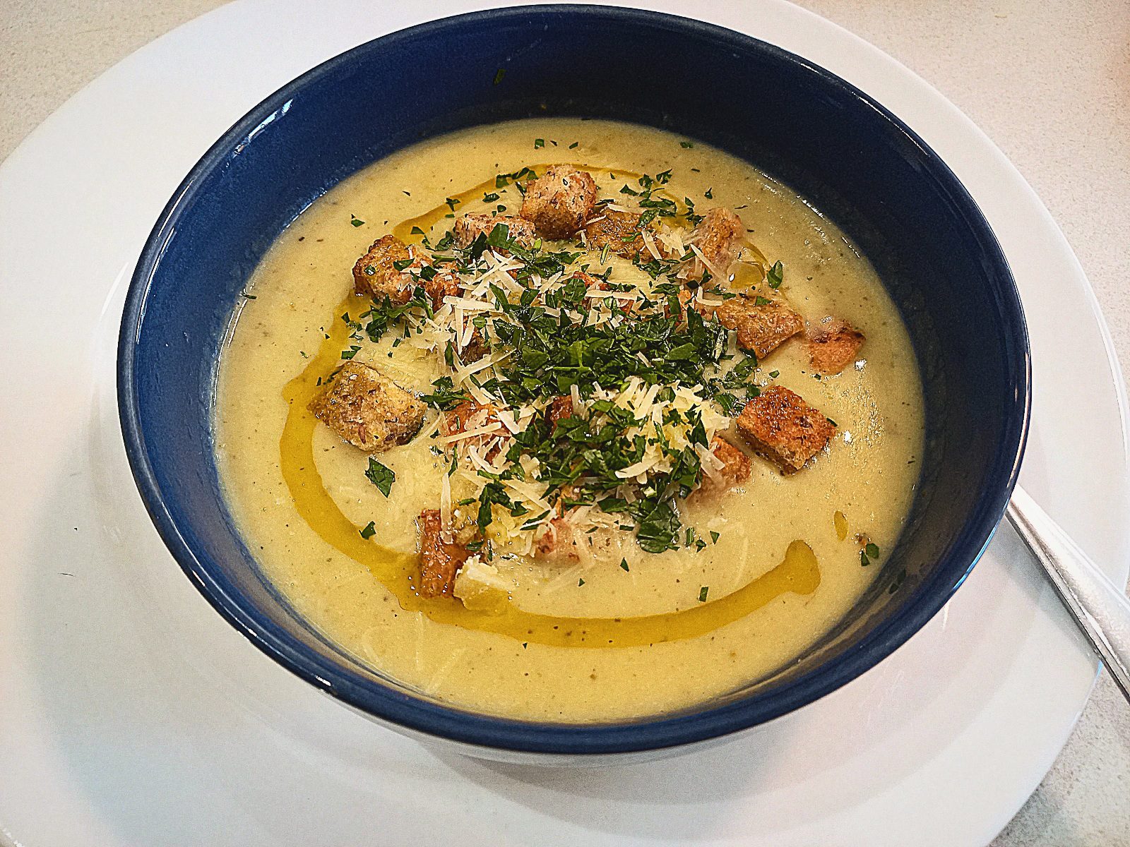 Roasted garlic cream soup topped with croutons, grated Parmesan cheese, chopped parsley and a drizzle of truffle oil. In a blue bowl on a white plate