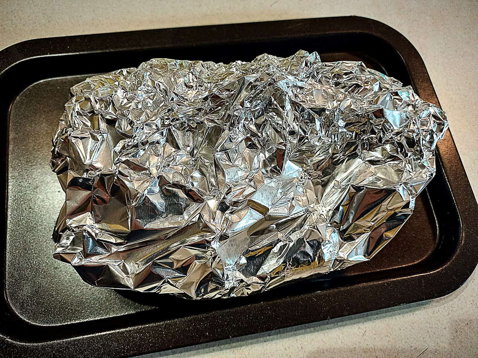 A tin foil parcel placed on a black oven tray sitting on a cream colored bench top