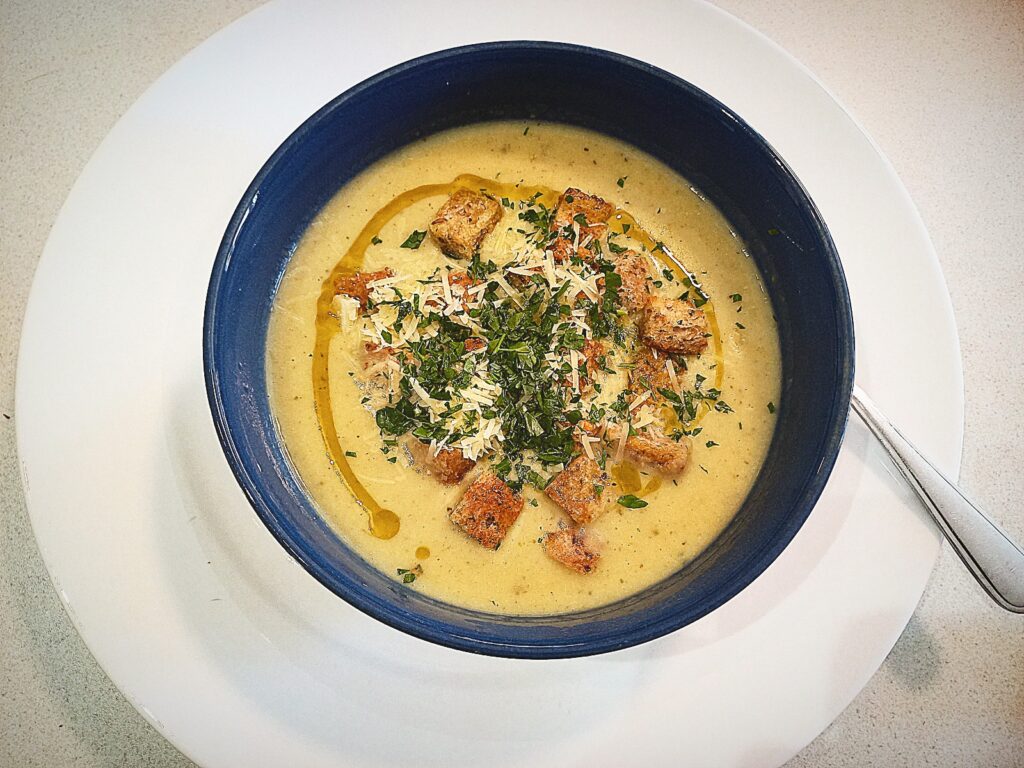 Roasted garlic cream soup topped with croutons, grated Parmesan cheese, chopped parsley and a drizzle of truffle oil. In a blue bowl on a white plate with a silver spoon