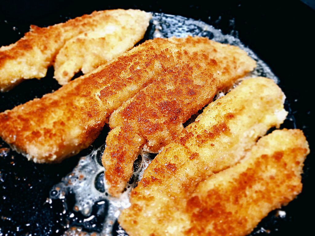 Three crumbed fish fillets frying golden brown in a cast iron skillet pan