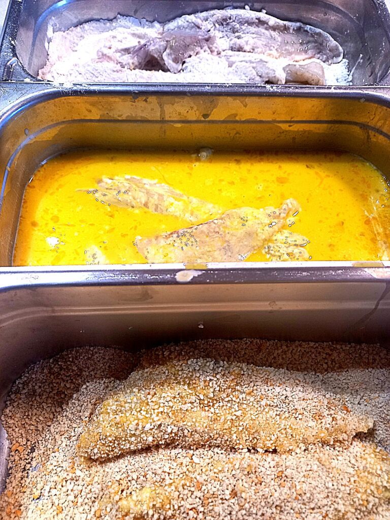 Three deep trays next to each other, one with flour, the second with egg wash, the third with bread crumbs, all of them with a fish fillet in it