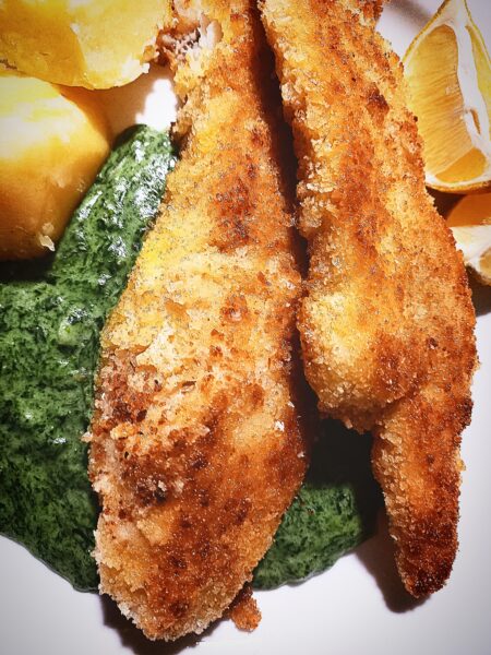 On a white plate are two pieces of pan fried fish on top of creamed nettle sauce. Two boiled potatoes and two lemon wedges next to it.