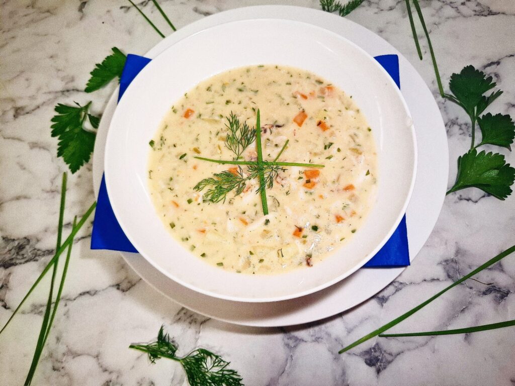 Crab chowder served in a white soup bowl on a serving plate with a blue square napkin. The soup bowl is placed on a marbled stone top with parsley leaves and chives around it.
