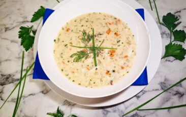 Crab chowder served in a white soup bowl on a serving plate with a blue square napkin. The soup bowl is placed on a marbled stone top with parsley leaves and chives around it.