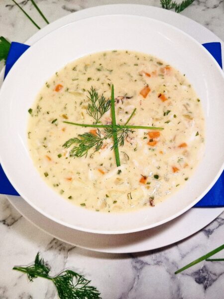 Crab chowder served in a white soup bowl on a serving plate with a blue square napkin. The soup bowl is placed on a marbled stone top with parsley leaves and chives around it.