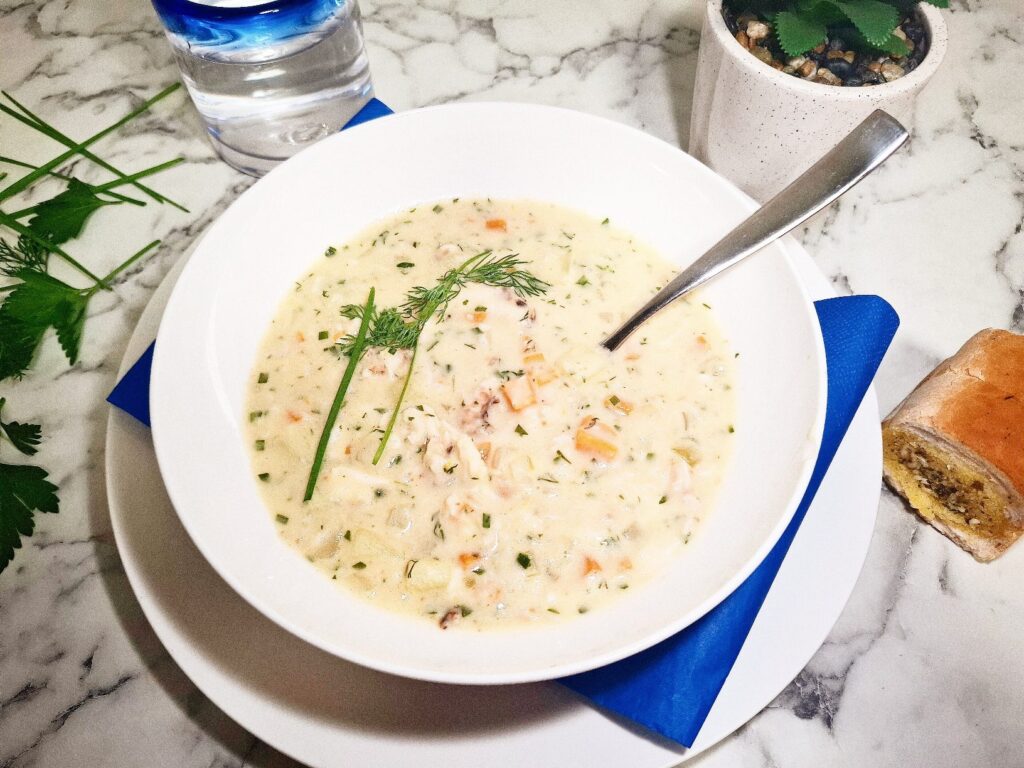 Crab chowder served in a white soup bowl with a spoon in it on a serving plate with a blue square napkin. The soup bowl is placed on a marbled stone top with parsley leaves and chives next to it as well as a drinking glass with blue ring, a small pot plant and a small bread roll.