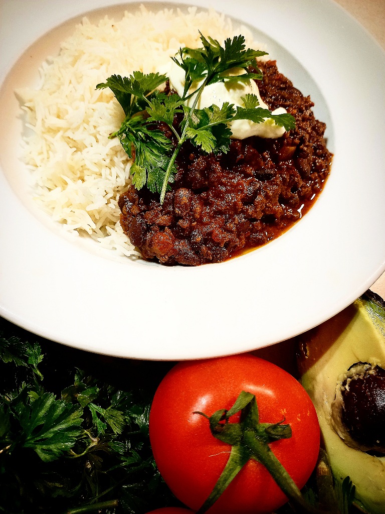 Black bean and venison chili with rice in a white bowl. Coriander leaves, a tomato and half an avocado in front of the plate