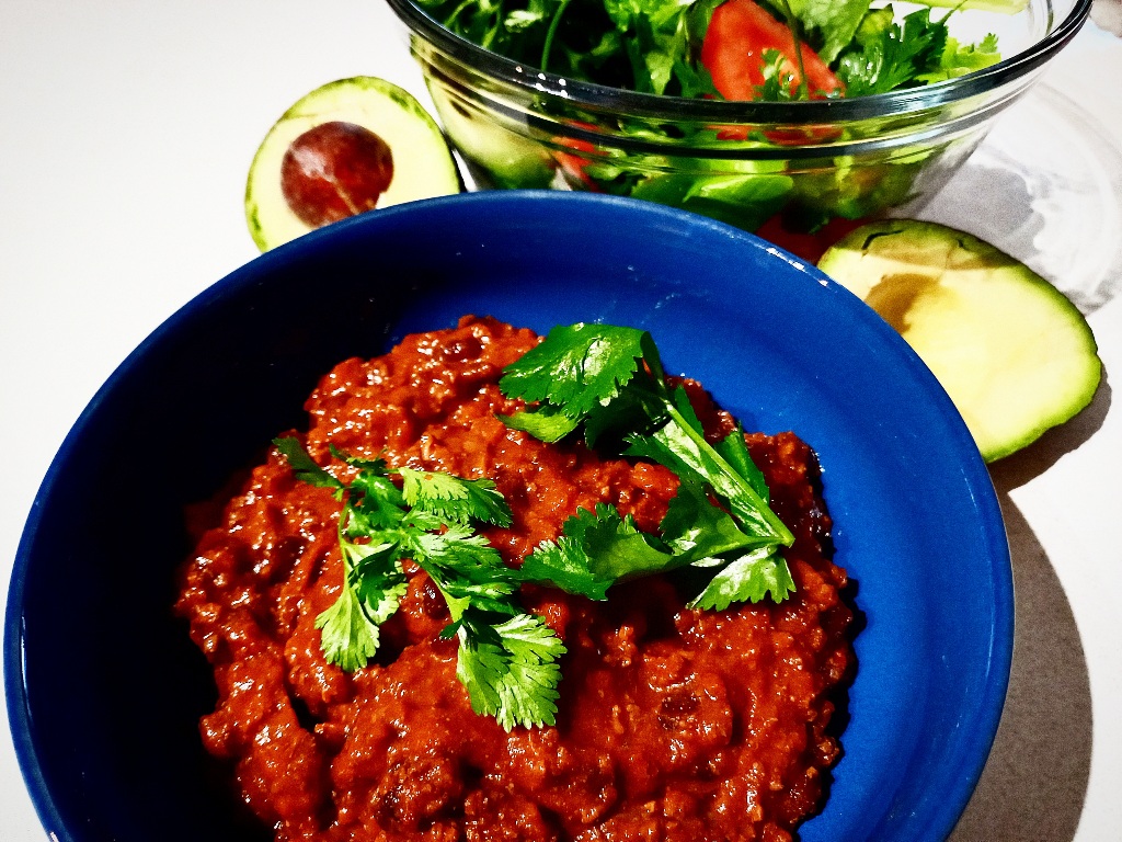 Venison chili with coriander leaves on top in a blue bowl. A bowl of salad and two avocado halves behind it.