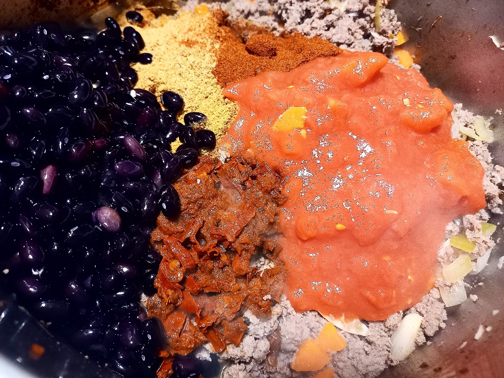 Ground venison cooking in a pot. Black beans, crushed tomatoes and spices sitting on top ready to be mixed in.