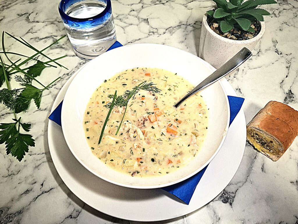 Crab chowder served in a white soup bowl with a spoon in it on a serving plate with a blue square napkin. The soup bowl is placed on a marbled stone top with parsley leaves and chives next to it as well as a drinking glass with blue ring, a small pot plant and a small bread roll.