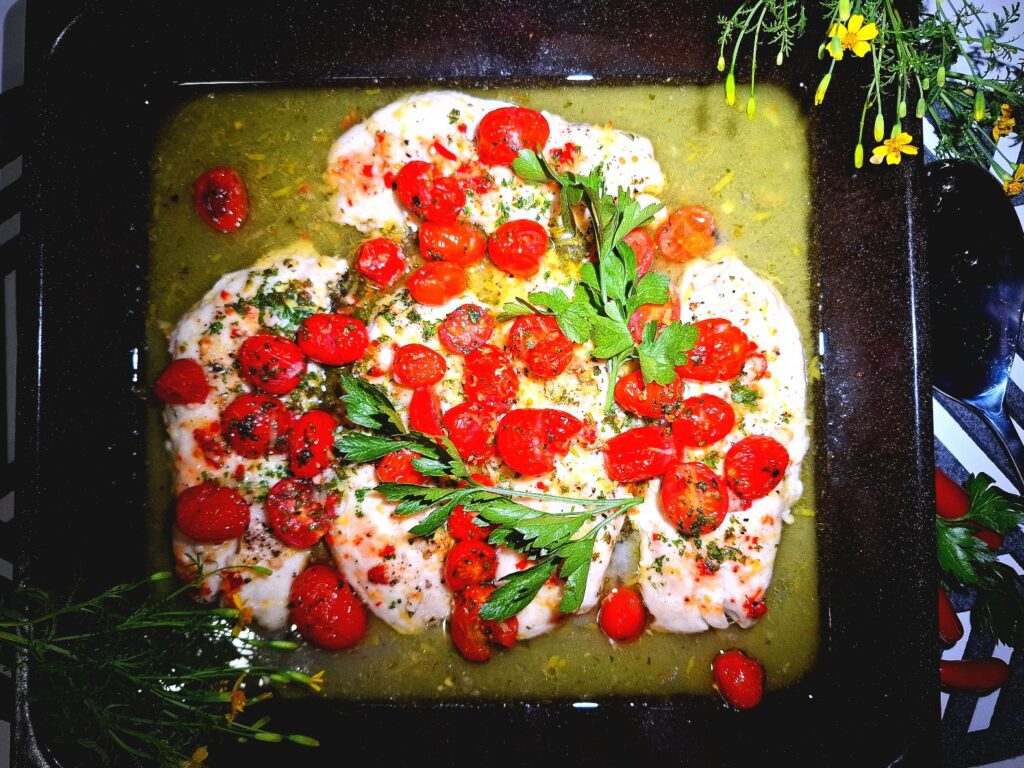 4 Baked fish fillets in an oven tray topped with tomato garlic and parsley. Silver serving spoon, garnished with yellow flowers.