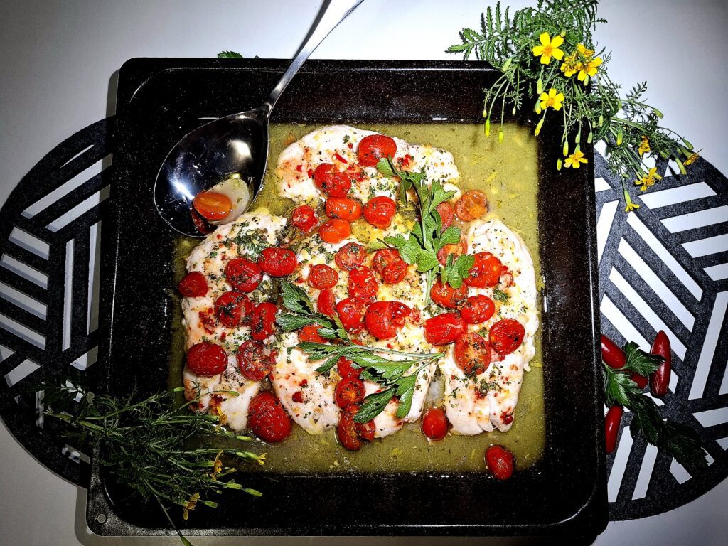 4 Baked fish fillets in an oven tray topped with tomato garlic and parsley. On a round place mat on a white table. Silver serving spoon, garnished with yellow flowers.