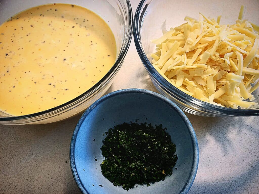 Three bowls on a bench top. One glass bowl with egg and cream mixture, another glass bowl with grated cheese. The third one a small ceramic one with chopped parsley.