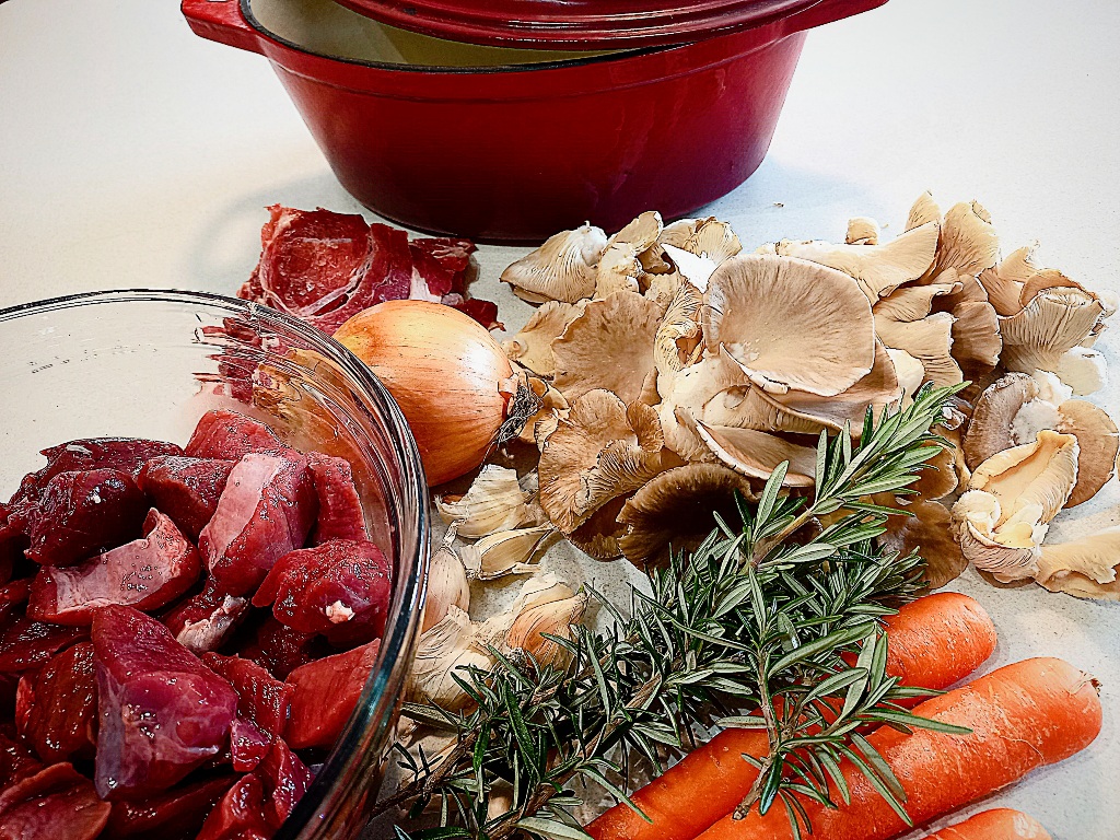 All the ingredients for venison stew on a white kitchen bench. A red dutch oven in the back ground
