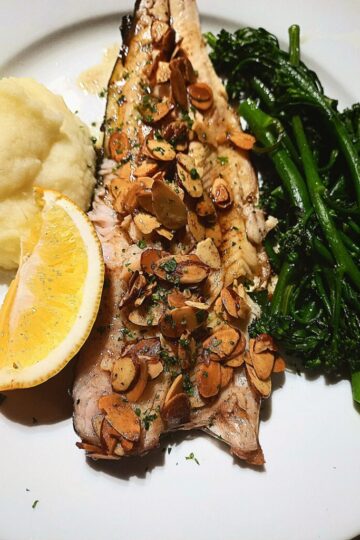 A Trout fillet on a white plate topped with almondine sauce. Mash potato and a lemon wedge on one side and steamed broccolini on the other side of the fillet