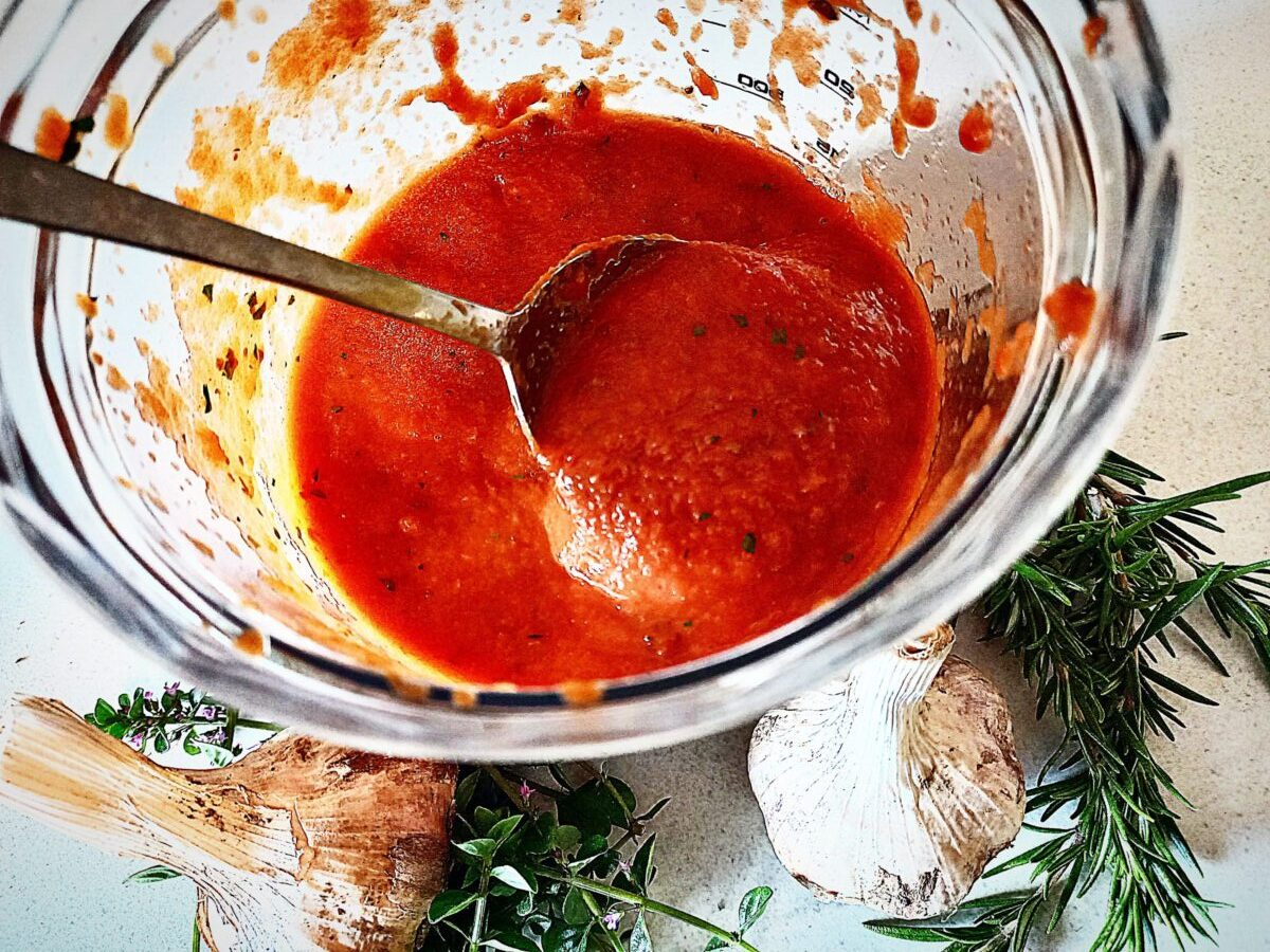 Pizza sauce in a clear blender base and a spoon in it. Two heads of garlic and fresh herbs in front of it