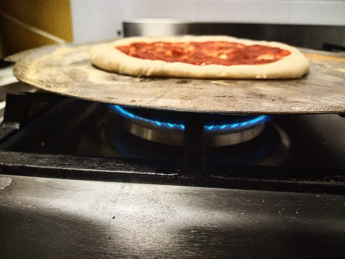Raw pizza base topped with pizza sauce cooking on a gas burner.