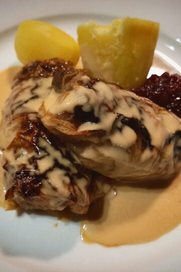 Two Swedish cabbage rolls on a white plate with two boiled potatoes, creamy sauce and lingon berry jam