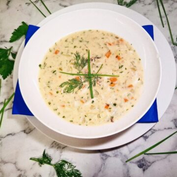 Crab chowder served in a white soup bowl on a serving plate with a blue square napkin. The soup bowl is placed on a marbled stone top with parsley leaves and chives around it.