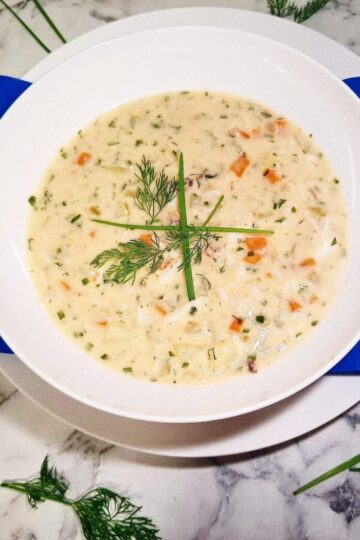 Crab chowder served in a white soup bowl on a serving plate with a blue square napkin. The soup bowl is placed on a marbled stone top with parsley leaves and chives around it.