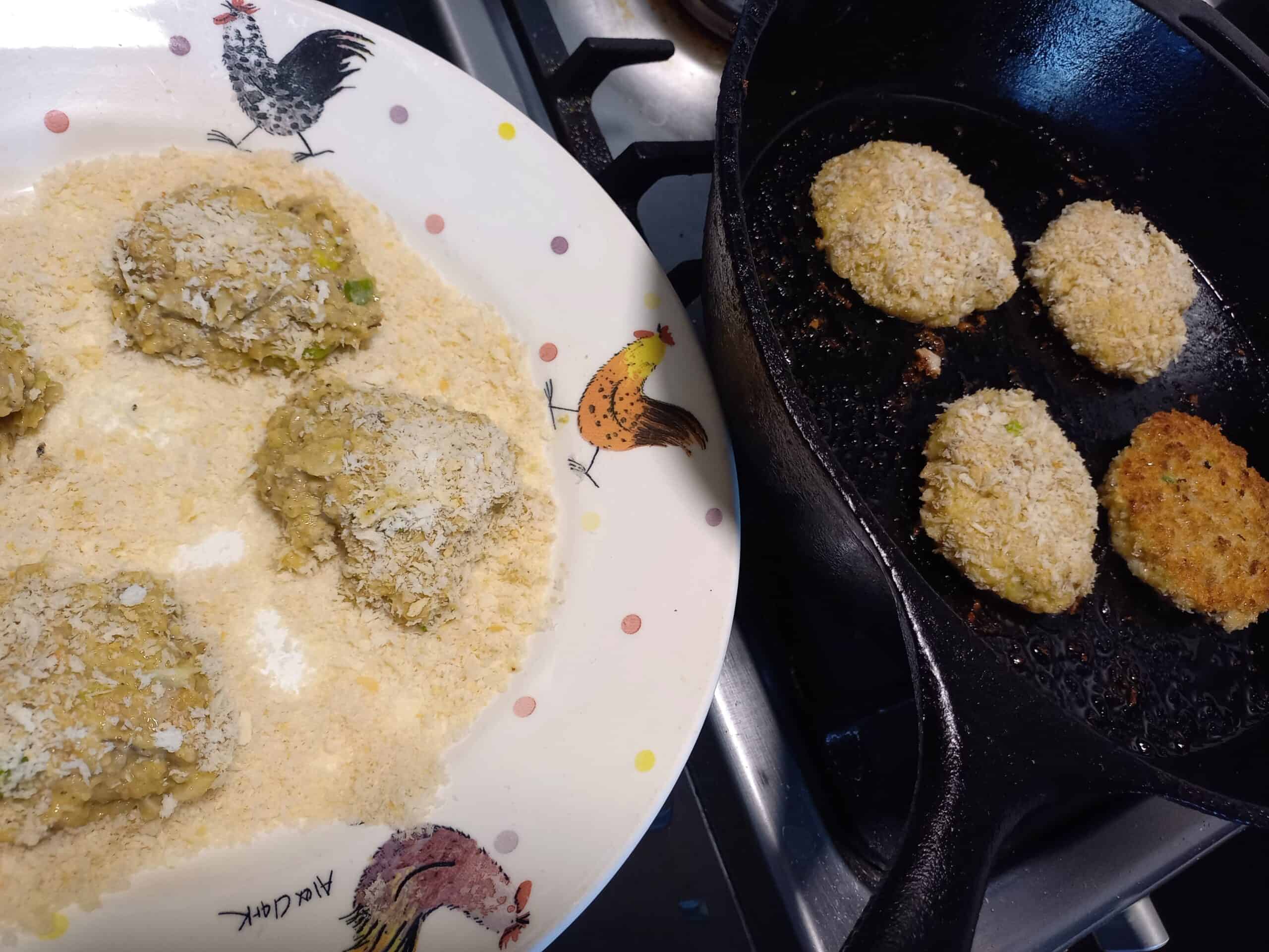 frying clam fritters in cast iron skillet frying pan, a plate with raw fritters rolled in breadcrumbs next to it.