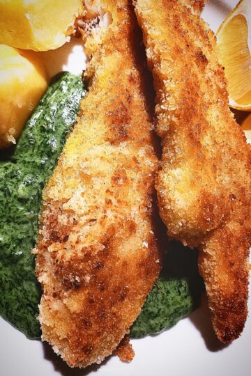 On a white plate are two pieces of pan fried fish on top of creamed nettle sauce. Two boiled potatoes and two lemon wedges next to it.