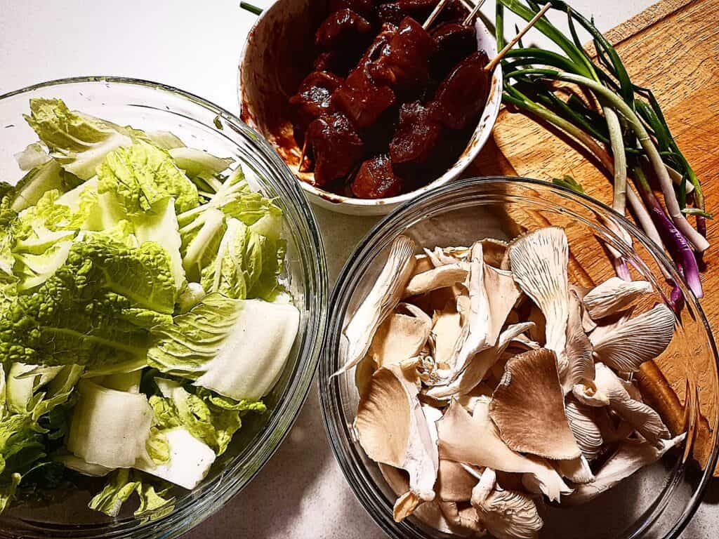 Cut savoy cabbage in a glass bowl, oyster mushrooms tared in another glass bowl. Marinated diced duck breast in a small white bow. A few spring onions laying on a wooden chopping board next to it