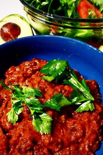 Venison chili with coriander leaves on top in a blue bowl. A bowl of salad and two avocado halves behind it.