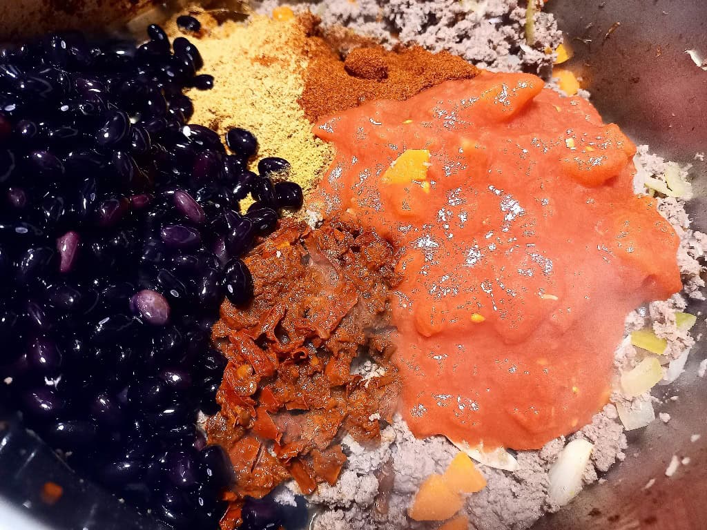 Ground venison cooking in a pot. Black beans, crushed tomatoes and spices sitting on top ready to be mixed in.