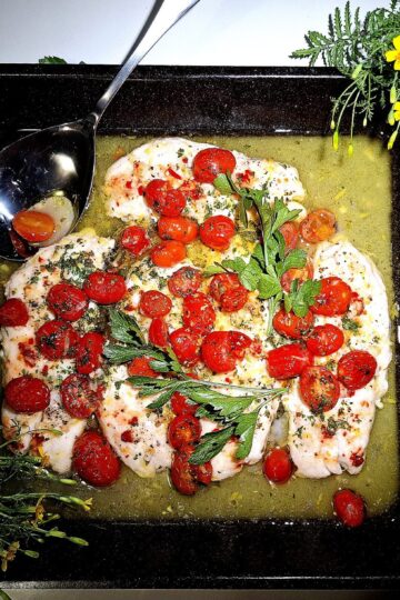 4 Baked fish fillets in an oven tray topped with tomato garlic and parsley. On a round place mat on a white table. Silver serving spoon, garnished with yellow flowers.