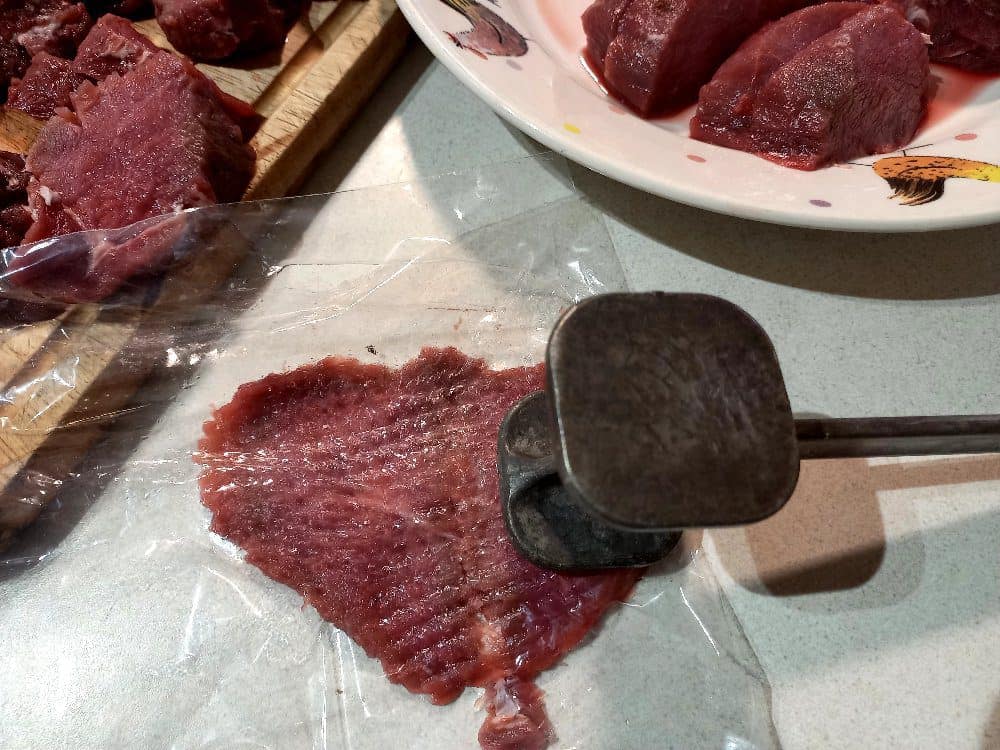 Venison slices on a plate, pounding meat with a hammer and a chopping board with meat slices on it