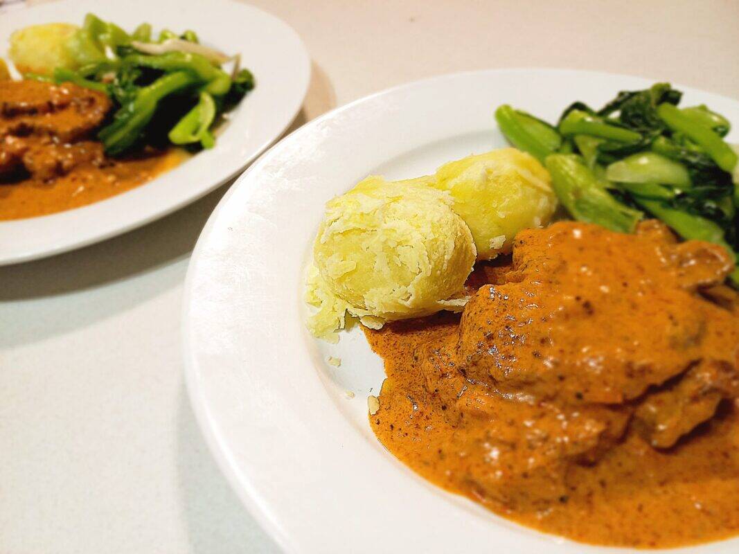two plates of pork pepper steak in a tomato cream sauce with boiled potatoes and steamed vegetables on a white kitchen bench