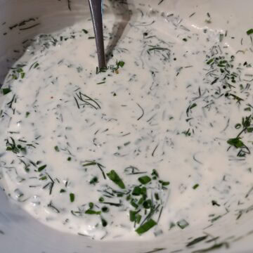 A close up image of sour cream lemon and herb dressing with in a white bowl and a spoon in it