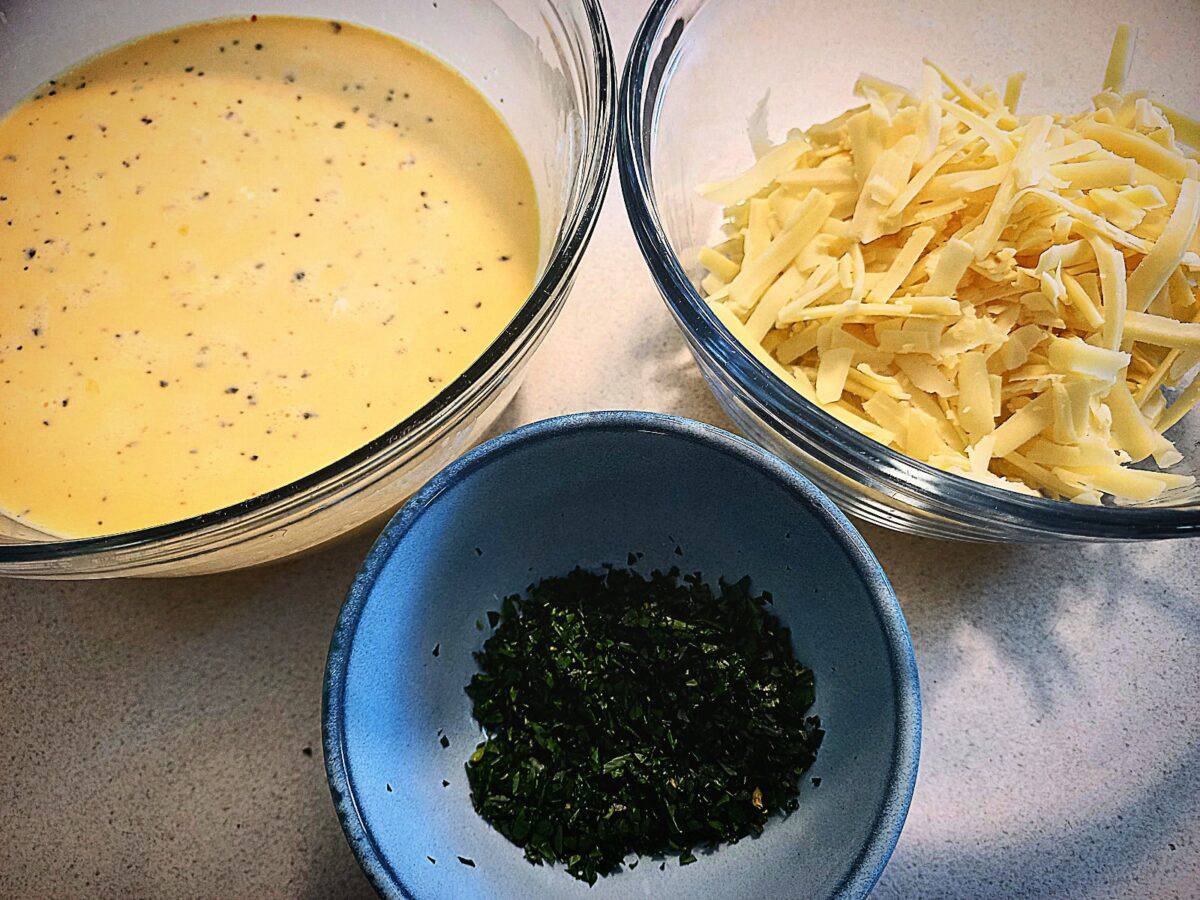 Three bowls on a bench top. One glass bowl with egg and cream mixture, another glass bowl with grated cheese. The third one a small ceramic one with chopped parsley.
