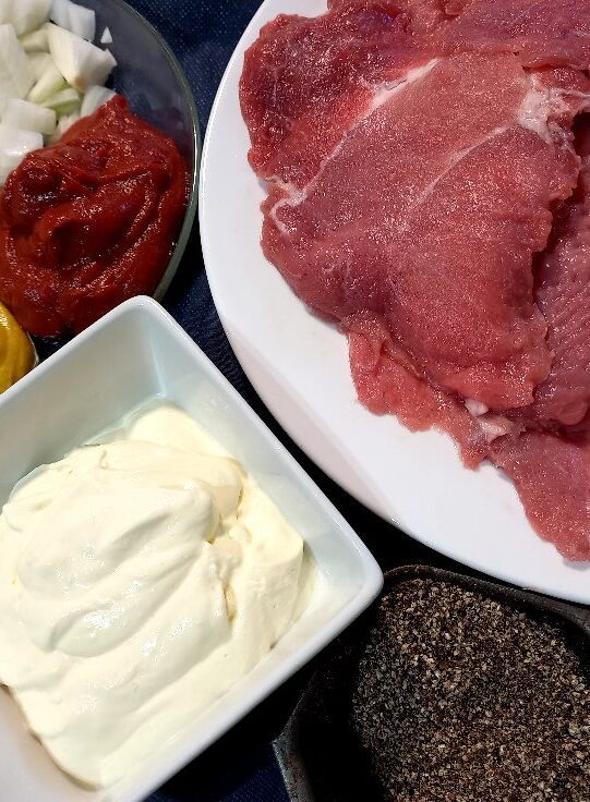 Thin slices of pork meat on a plate, a bowl of diced onion, tomato paste and mustard, a small ramekin with cream and a ramekin with coarsely ground pepper