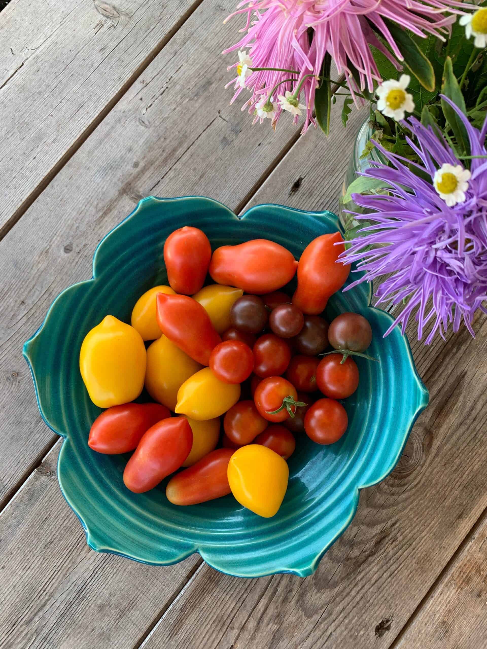 small colorful tomatoes in a blue bowl on a wooden table and a flower vase next to it