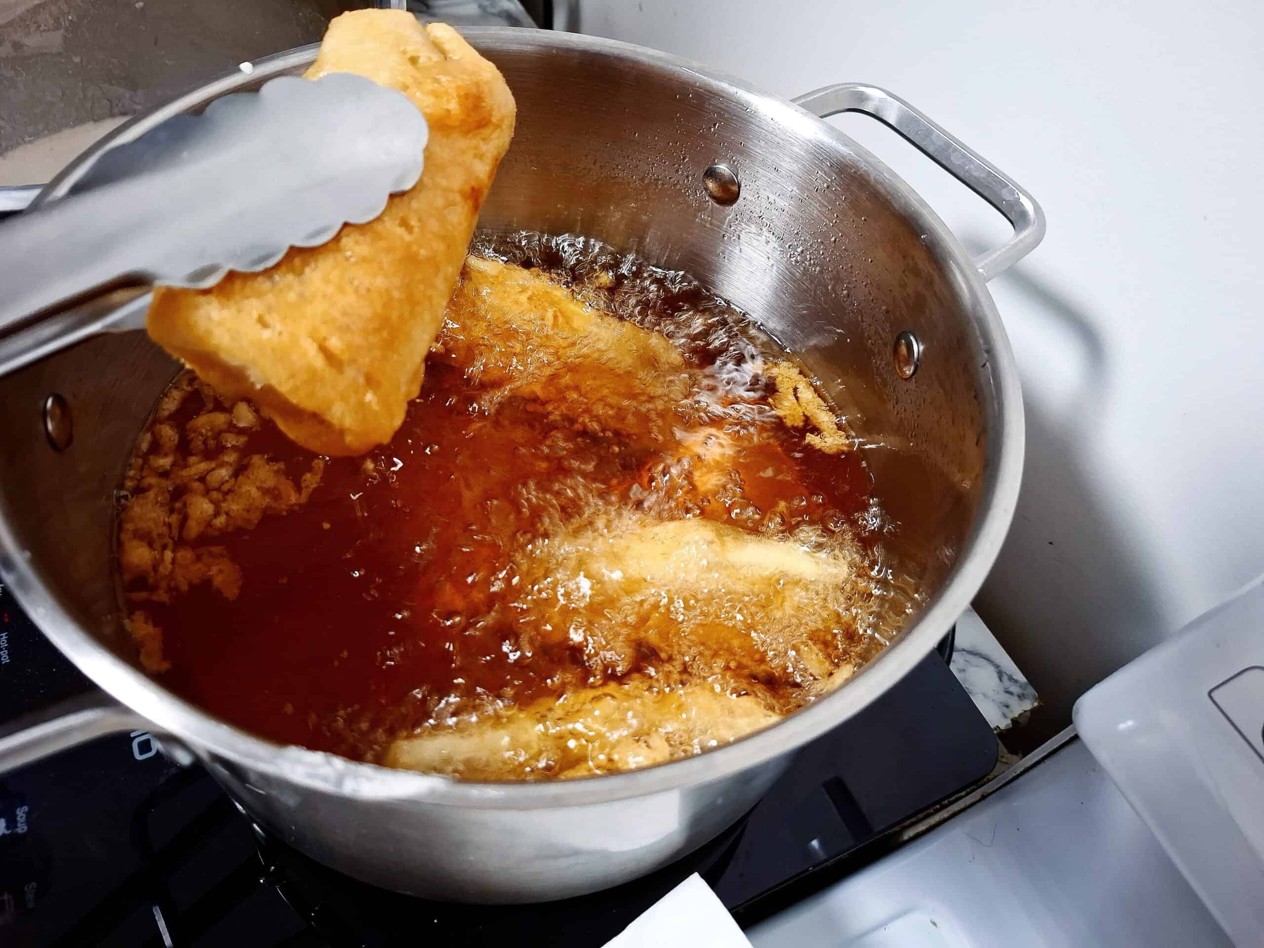 3 tempura fish pieces cooking in hot oil in a saucepan and another piece is being picked up by kitchen tongs